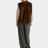 Person wearing a brown fur vest over a beige shirt with gray pants on a white background